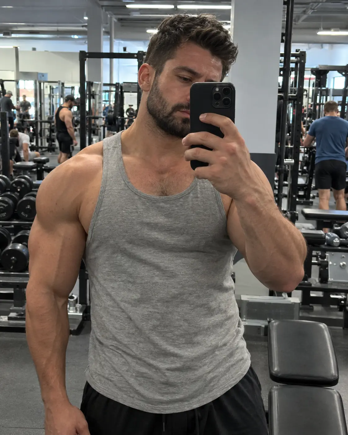 A muscular man with a trimmed beard takes a mirror selfie in a busy gym, his phone partly covering his face